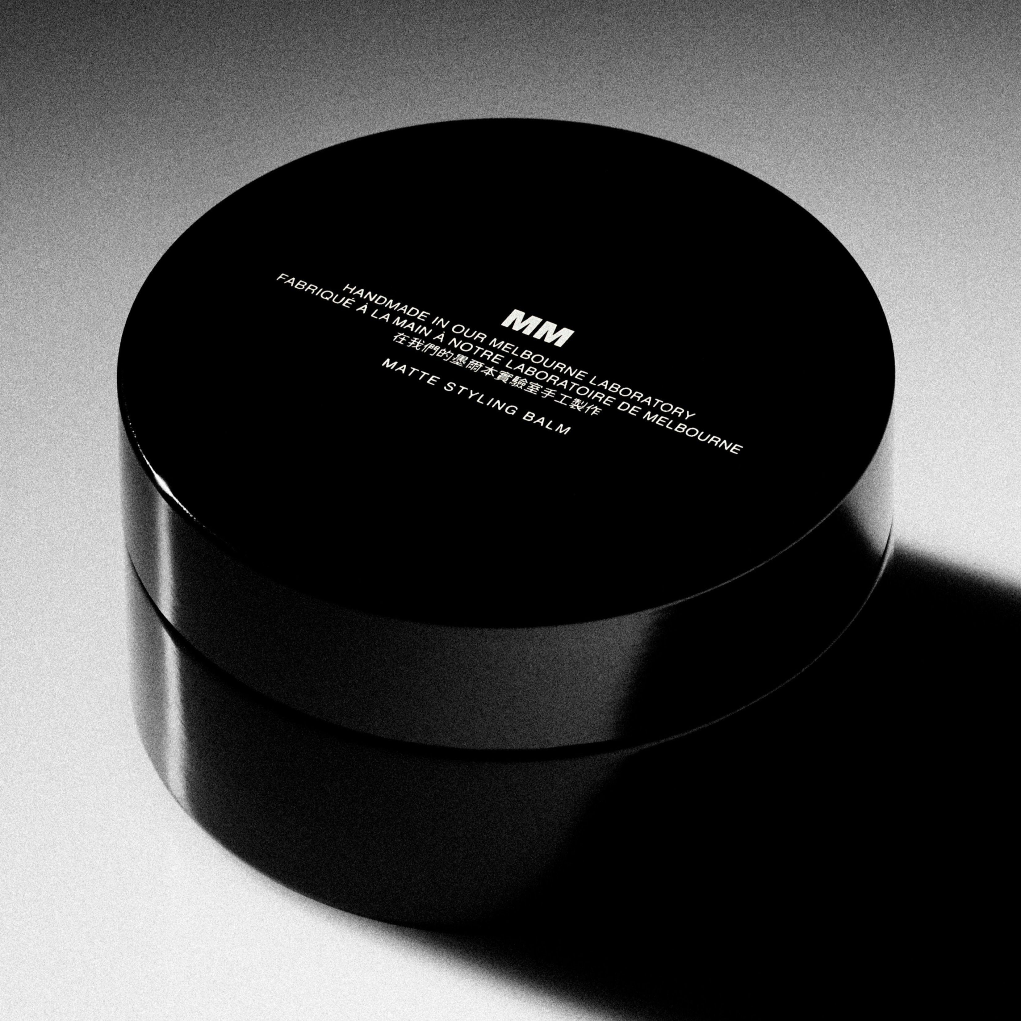 A black cylindrical container with a matte finish, labeled 'Morris Motley New MATTE STYLING BALM premium hair clay ' on a textured grey background